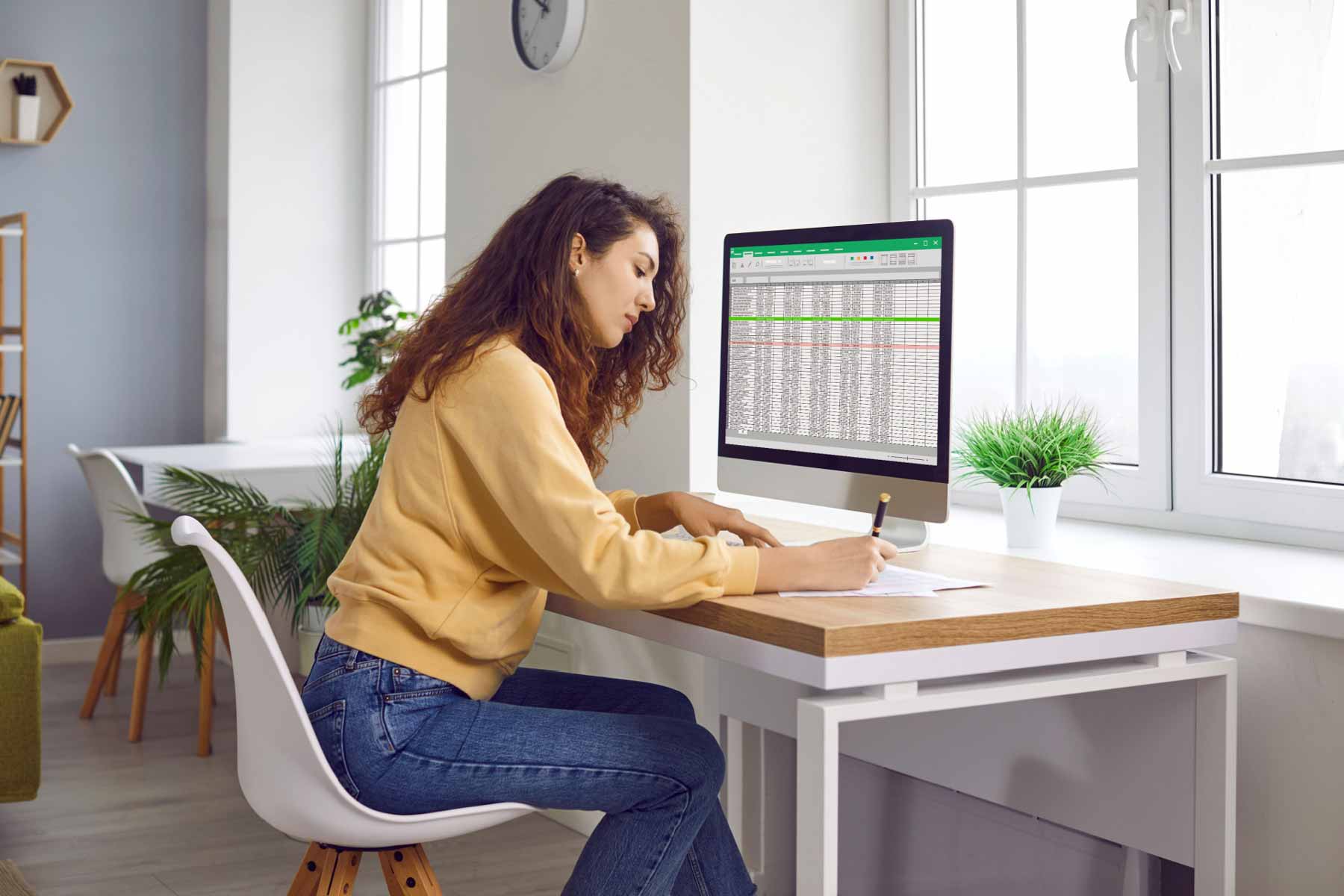 Young woman working at a modern desk, viewing spreadsheets on a large computer monitor and writing notes on paper in a bright office with plants